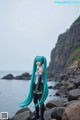 A woman with long blue hair standing on a rocky beach.